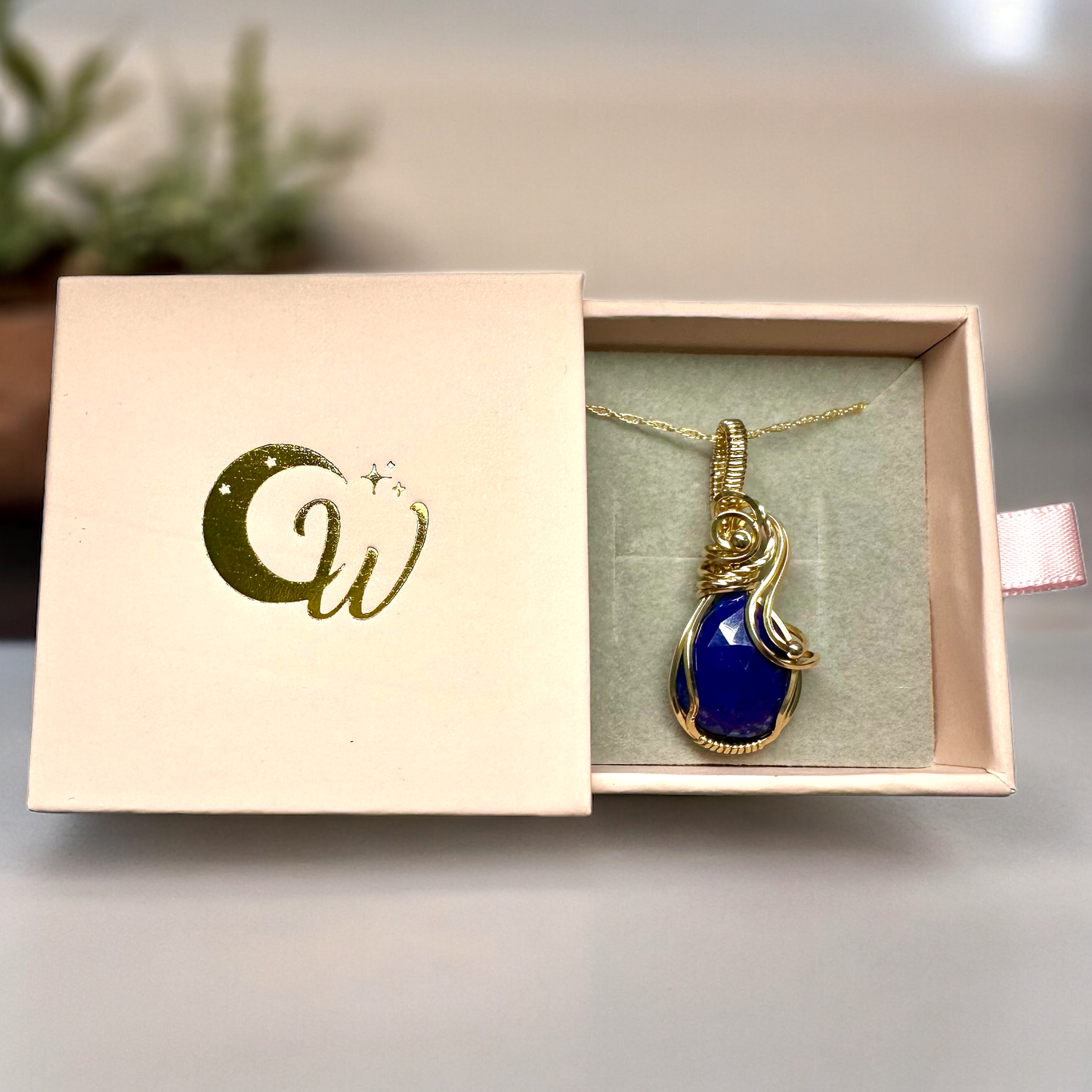 Faceted lapis lazuli teardrop necklace wrapped in 14K gold filled wire, presented in a light pink box with golden logo, symbolizing luxury and elegance.