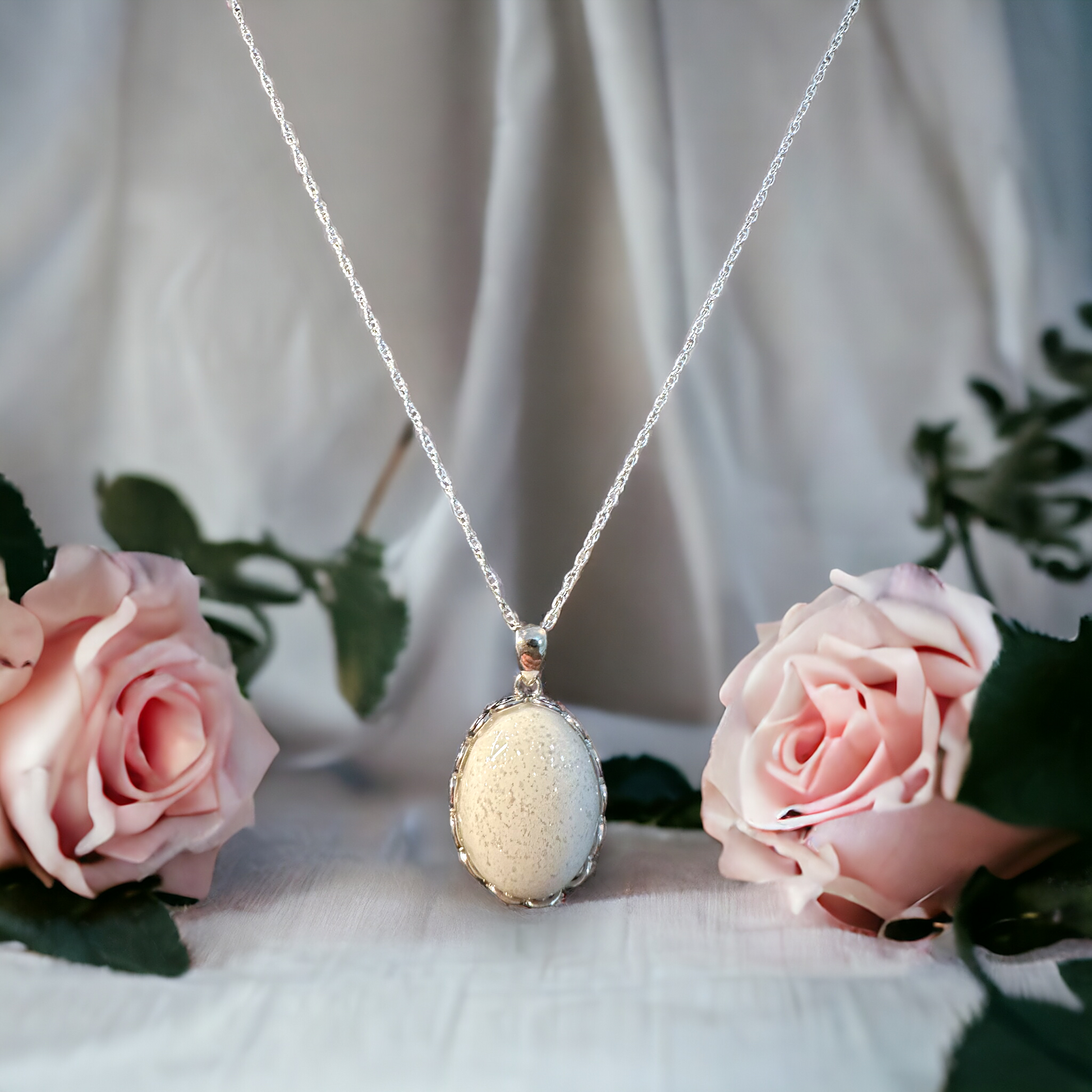 Eternal Grace sterling silver filigree necklace with a white breastmilk stone filled with crystal sparkles, elegantly hanging in front of flowers on a silver chain, highlighting the unique beauty of memorial jewelry.