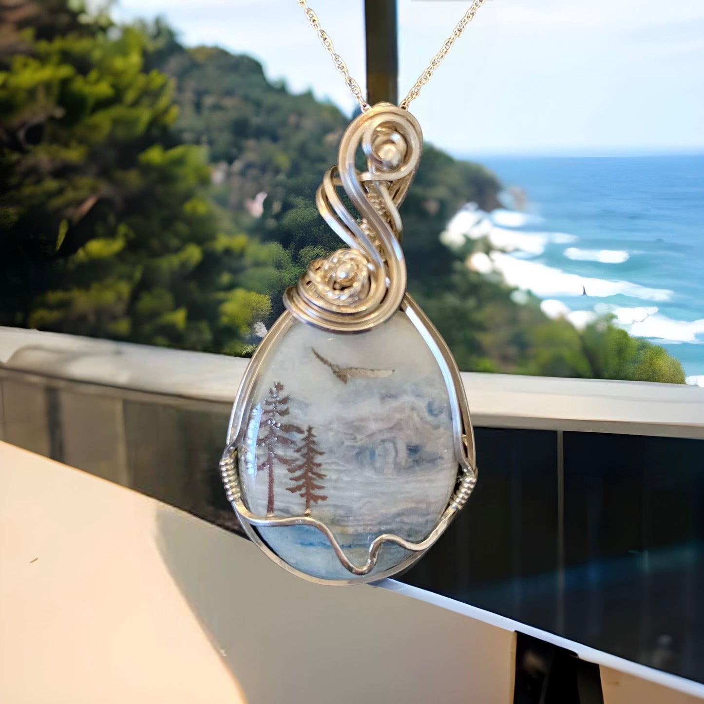 SSterling Silver and Blue Scheelite Beach Memorial Necklace hanging on a chain next to a window with a view of the ocean.